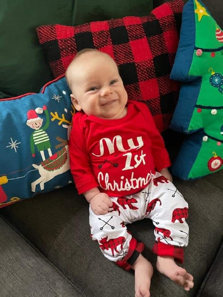 Smiling infant dressed in My First Christmas pajamas and propped against festive pillows