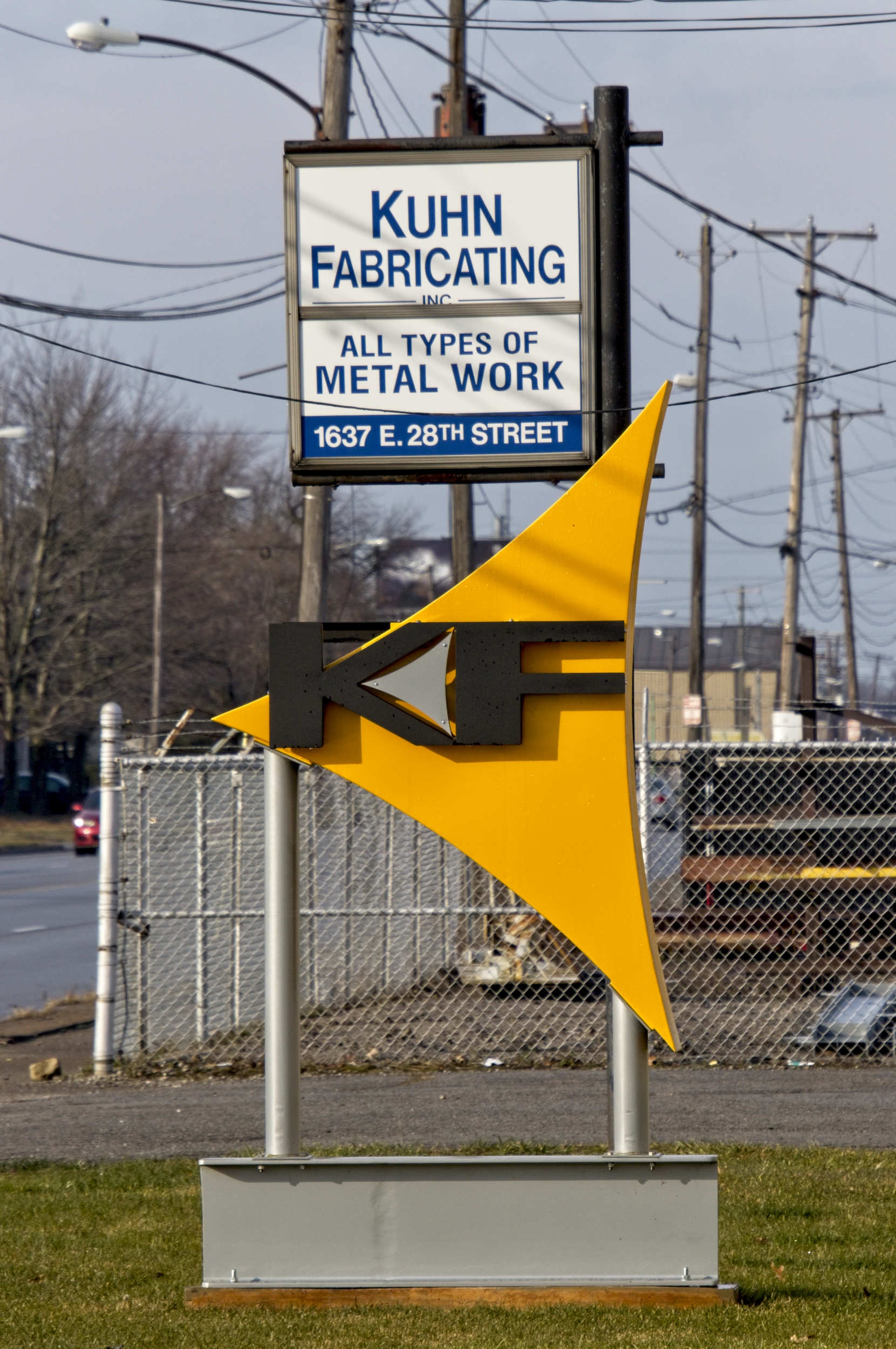 2 signs for a business. One is square and has the name of the business, what they do, and the address in blue letters on a white background. The other sign is the company logo: the letters K and F in black with a small gray triangular accent between them, all on a yellow triangular background. This is mounted to two metal posts which are mounted to the top of a section of steel beam.