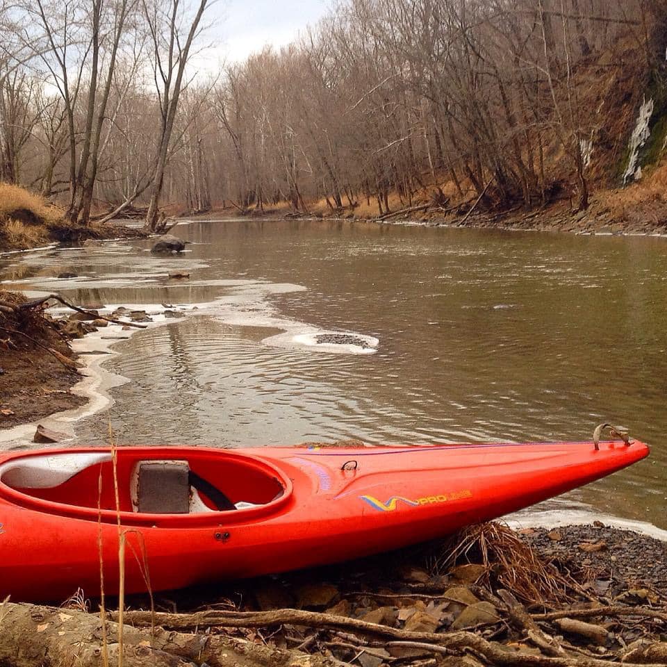 Red kayak sitting on the riverbank. The water is muddy brown, the trees are bare, and there is some ice along the shoreline.