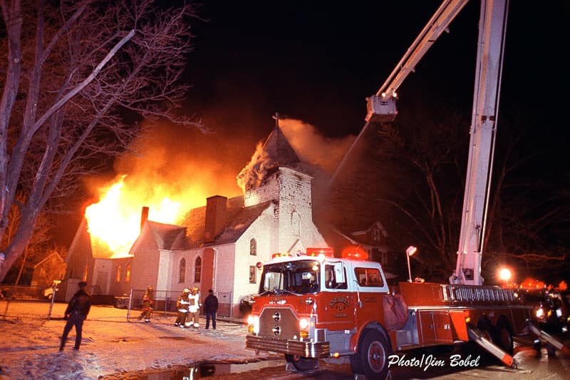 Large old church on fire in the background, large fire truck with aerial ladder extended in foreground