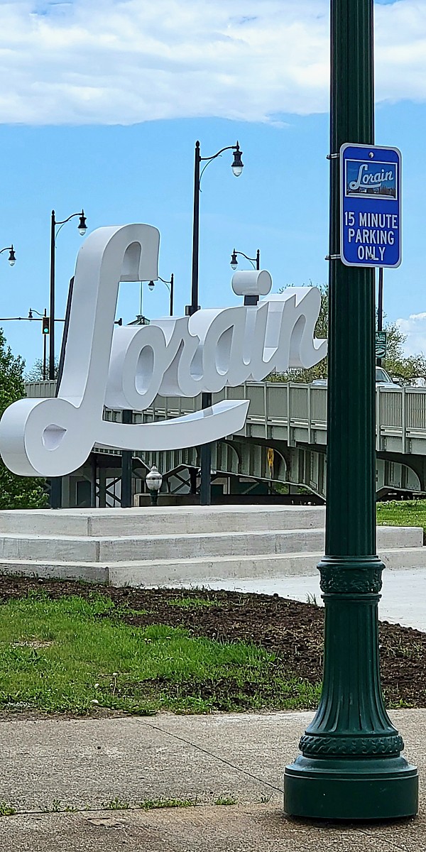 Photo of Script Lorain sign with Bascule Bridge in the background. In the foreground is a light pole with an affixed sign that shows a photo of the Script Lorain sign and "15 Minute Parking Only". This is in a dedicated right turn lane for Black River Landing.