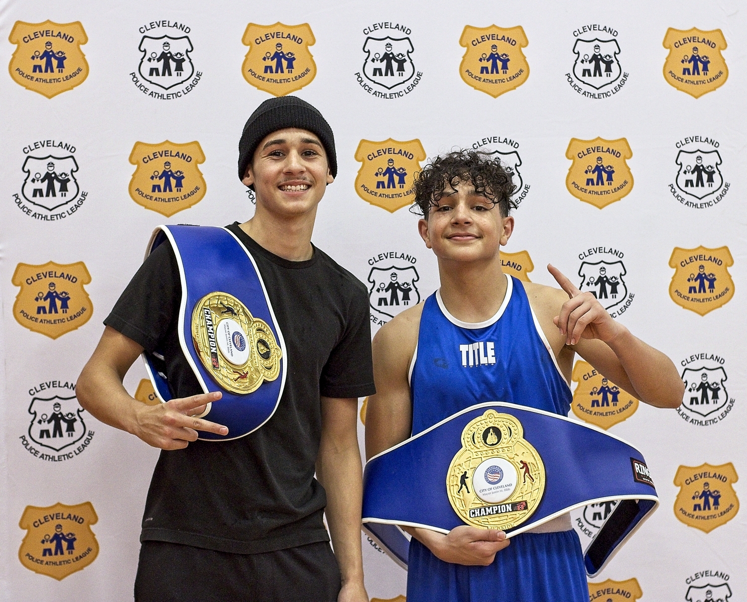 Two young men display their Champion belts while standing in front of the Cleveland Police Athletic League logo