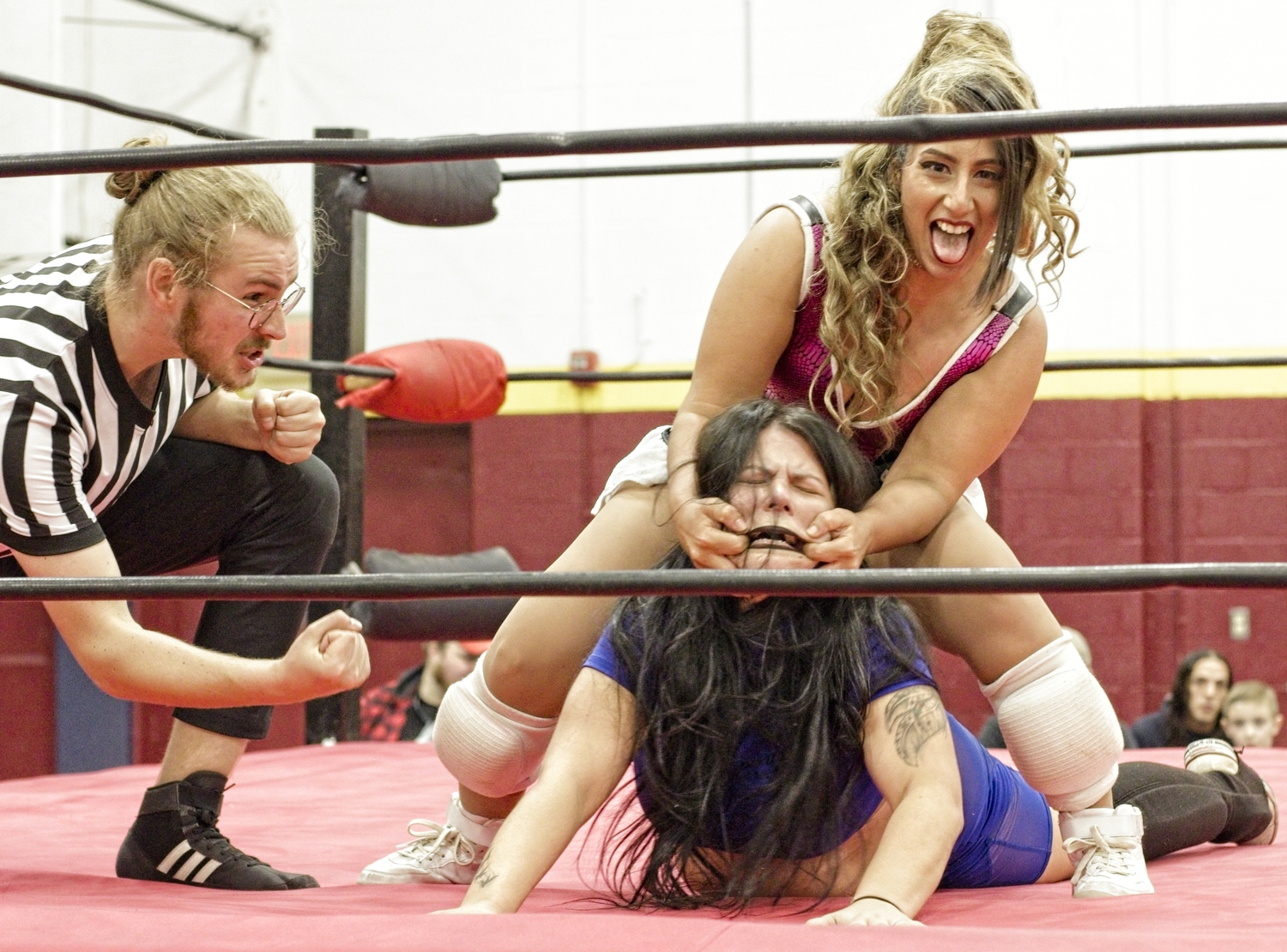 Local amateur wrestling match featuring female wrestlers. Male referee on the left is crouched down to keep a close eye on the action. Wrestler A is grinning wickedly while standing over Wrestler B who is laying on her stomach. Wrestler A has hooked her fingers into the corners of Wrestler B's mouth, simultaneously pulling her head up and her mouth into a grimace.