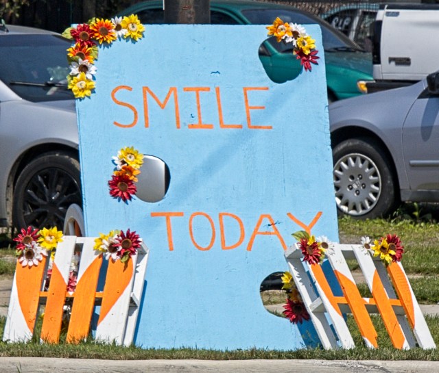 Blue sign with orange letters reads "Smile Today". It is propped against a utility pole on a street corner in South Lorain. At the foot of the sign, are two small white pallets painted with orange hearts. Everything is adorned with colorful flowers.