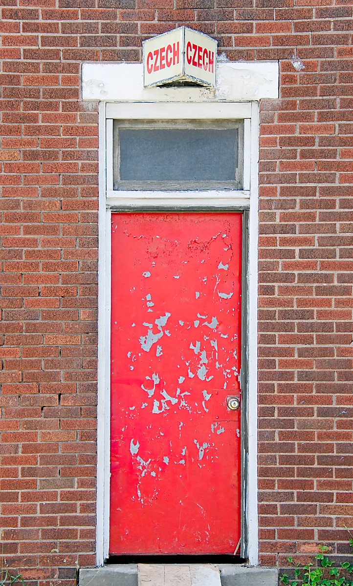 Beat up red door in a red brick building. There's a small window above the door and a 2 sided white sign with red lettering above that that reads Czech Czech. Used to be a side entrance for the long closed Czech Grill.